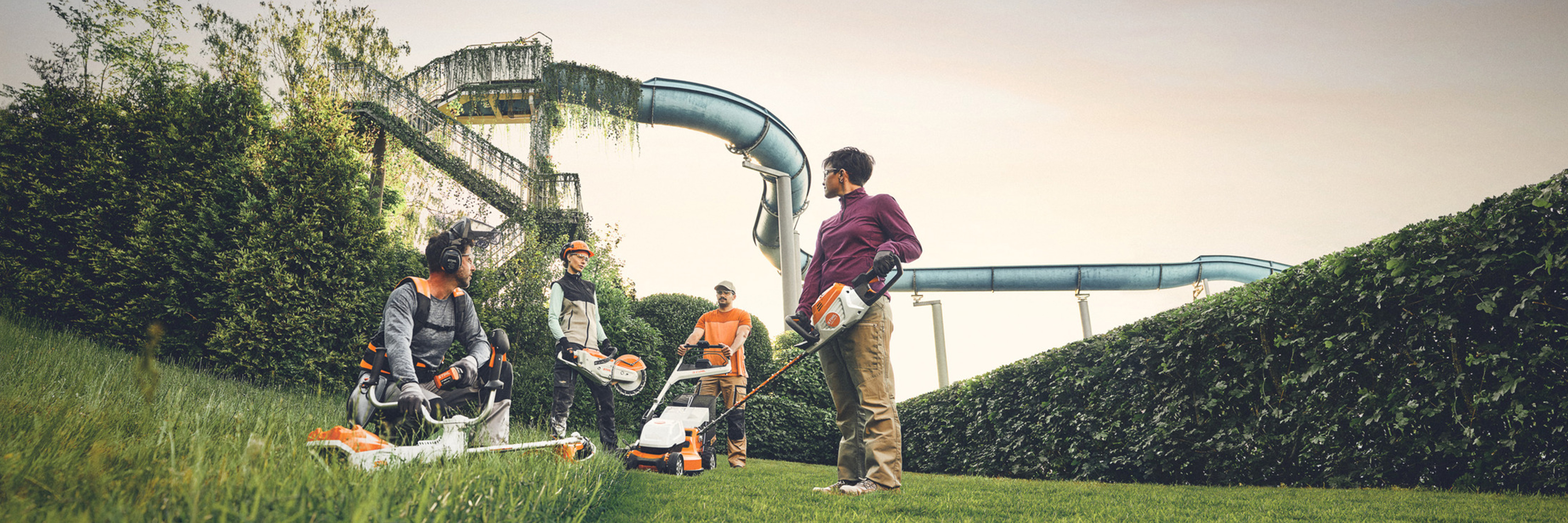 Grup njerëzish punojnë me vegla kopshtarie STIHL në një zonë të gjelbër me një gardh të madh dhe rrëshqitëse uji në sfond. Të pajisura me motokosa, kositëse bari dhe pajisje mbrojtëse.