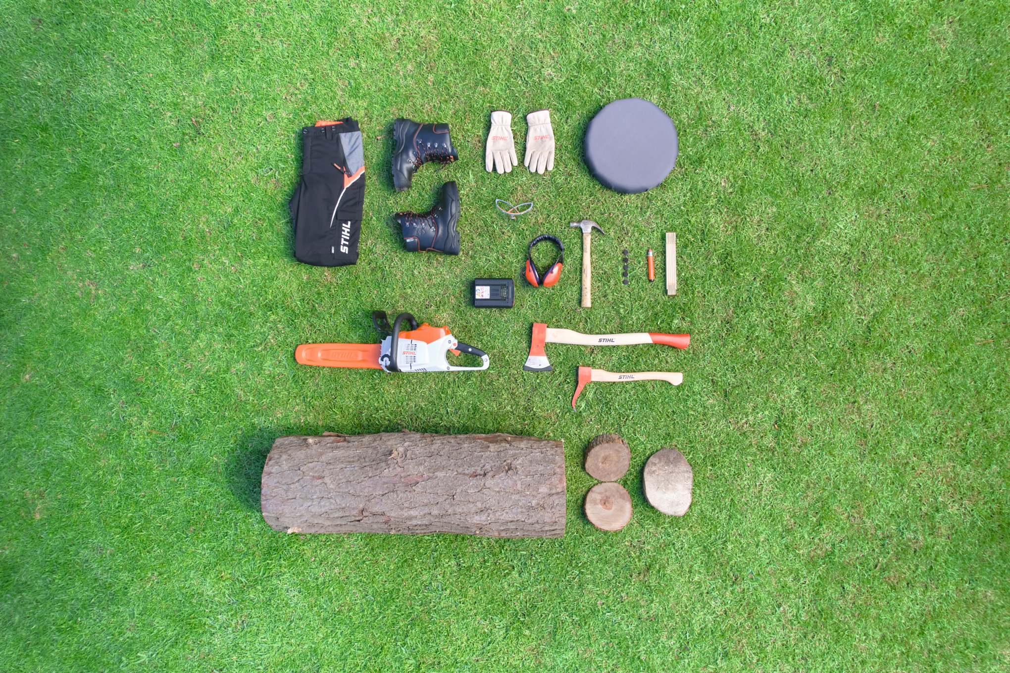Pajisje dhe mjete STIHL për punë me sharrë motorike, të vendosura mbi bar: pantallona mbrojtëse, çizme, doreza, syze mbrojtëse, mbrojtëse për dëgjimin, bateri, sharrë motorike, sëpata, çekiç, pyka dhe një trung druri.