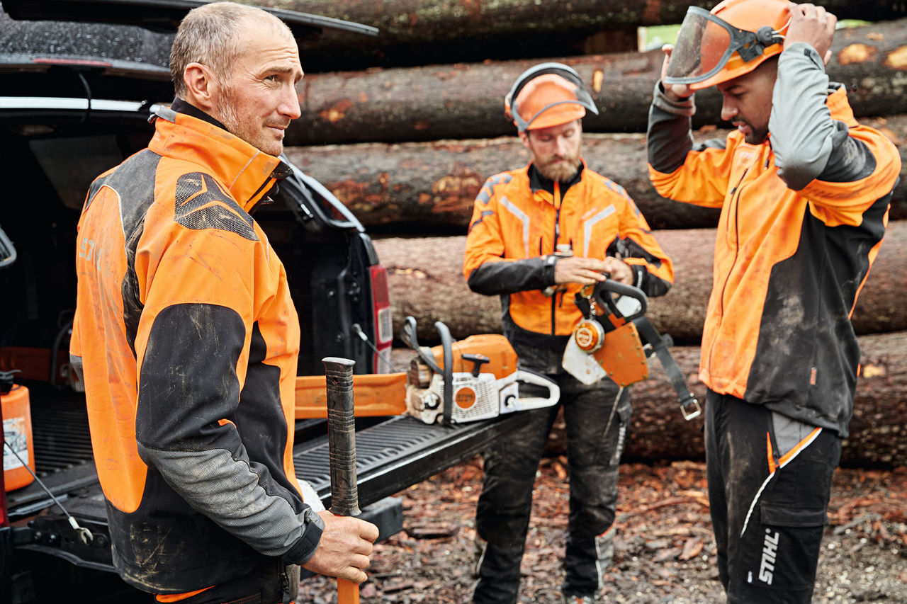 Tre burra të veshur me pajisje mbrojtëse para një automjeti, me bagazhin e hapur e në të të vendosura vegla elektrike pa kabllo STIHL