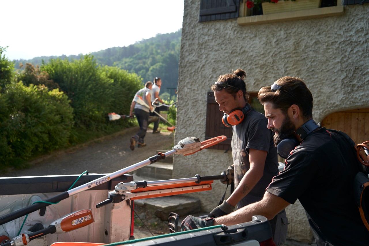 Dy kopshtarë peizazhesh shkarkojnë veglat me bateri STIHL nga një makinë, me punëtorë të tjerë në sfond 