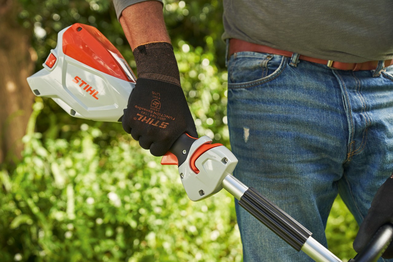 Një burrë që mban doreza mbrojtëse duke shtypur bllokuesin e këmbëzës së motokosës pa kabllo STIHL FSA 60 