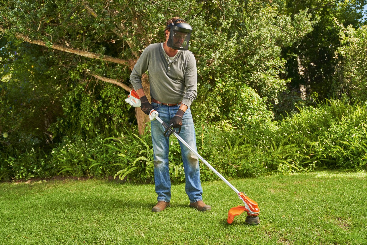 Një burrë që mban mbrojtje për fytyrë duke kositur barin në një kopsht me kositësen pa kabllo STIHL FSA 60