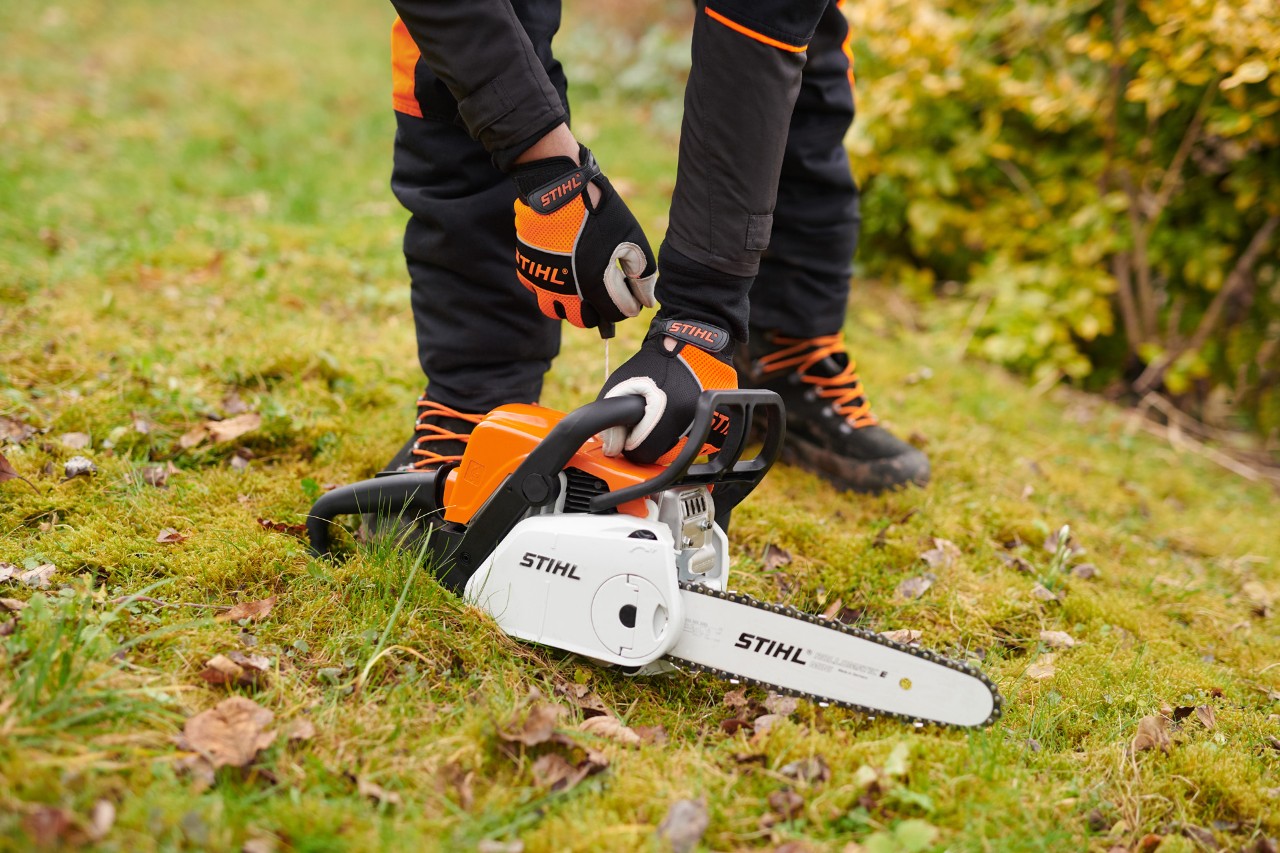 Motosharra STIHL MS 180 C-BE e shtrirë përtokë, me një person të veshur me doreza mbrojtëse që mban dorezën. 